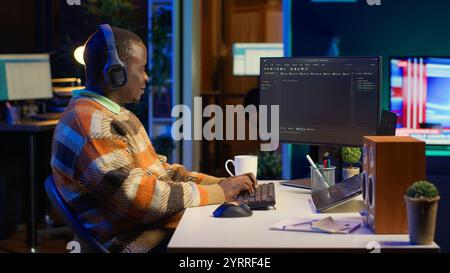 Black programmer remotely working in home office writing code on computer at his desk. Programming engineer IT expert in apartment typing lines of code at workstation, teleworking. Camera A. Stock Photo
