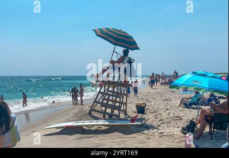 Robert Moses, New York, USA - 22 July 2023: A lifeguard sits on a tall chair under an umbrella as beachgoers enjoy the sunny day by the ocean. Stock Photo