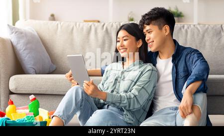 Loving asian couple with cleaning tools resting, using digital tablet Stock Photo