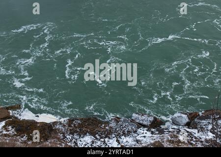 Close-up view of swirling river water with frothy patterns and a snowy, rocky shoreline in winter, Niagara Falls, Canada. Stock Photo