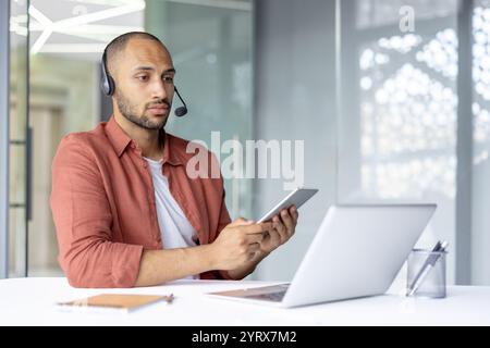 A young professional man is using a smartphone and laptop at his desk, wearing a headset. The scene suggests a remote work environment, highlighting technology use and communication. Stock Photo