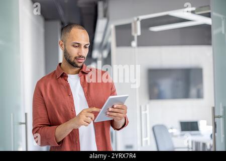 A man stands in a modern office, engaged with a tablet. He appears focused and professional, highlighting the use of technology in a workplace setting. Stock Photo