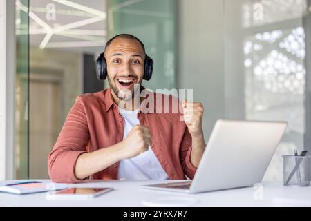 Young man enthusiastically cheers in office, celebrating success. He wears headphones and sits at a desk with a laptop. The setting conveys a sense of accomplishment and joy. Stock Photo