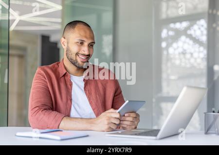 A smiling office worker interacts with a tablet and laptop in a contemporary workspace. The atmosphere is professional and innovative, highlighting the use of technology in a modern office setting. Stock Photo