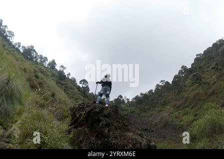 A lone hiker stands on a rocky outcrop at Volcano El Ajusco in Mexico City. Surrounded by lush green slopes and dense trees, the atmosphere is serene Stock Photo