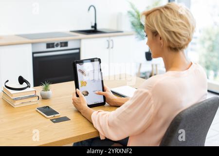Caucasian woman ordering food online using tablet in modern kitchen setting. She is reviewing menu items and prices on delivery website. Scene depicts convenience of online shopping. Stock Photo