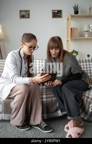 Serious doctor showing tablet screen to female patient, examination result, and giving consultation at home. practitioner visiting woman Stock Photo