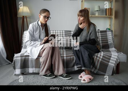 female doctor counseling an elderly woman at home. Writing notes on medical form at clipboard sitting on the sofa at home during visit. Stock Photo