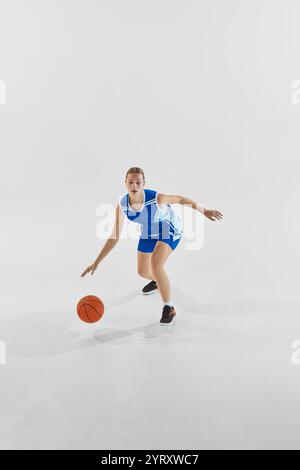 Full-length dynamic image of young teen girl, basketball player in blue uniform in motion with ball during game, training against white studio Stock Photo