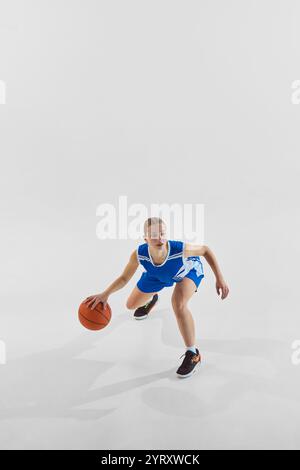 Full-length dynamic image of young teen girl, basketball player in blue uniform in motion with ball during game, training against white studio Stock Photo