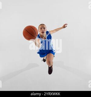 Slam dunk. Top view dynamic image of young focused teen girl in blue uniform, basketball player in motion, jumping with ball against white studio Stock Photo