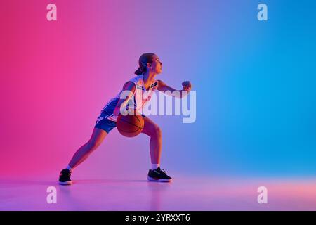 Dynamic image of young teen girl, basketball player in motion with ball, training, playing against gradient pink blue background in neon light Stock Photo