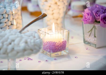 Lit candle centerpiece illuminating sugared almonds and purple roses at wedding reception Stock Photo