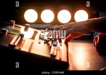 Close-up of a technician’s hands operating a sound mixing console, illuminated by stage lighting, emphasizing audio production at a live event. Stock Photo