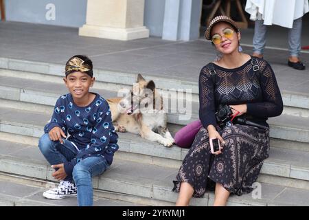 Bali, March 6 2020Young boy wearing a crown, smiling with a dog nearby, seated next to a woman in sunglasses and a patterned outfit, outdoors on stone Stock Photo