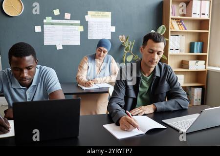 Medium shot of adult multiracial man concentrated on writing task sitting at desk in study group with other students attending language courses class Stock Photo