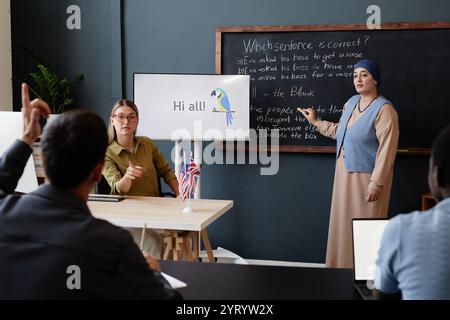 Adult Muslim woman standing at blackboard completing multiple choice task while classmate helping with hint explaining English grammar during lesson at language school Stock Photo