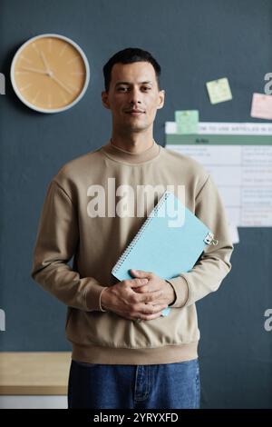 Vertical portrait shot of happy mixed raced male student posing with exercise book in hands while standing against blue wall in school classroom, copy space Stock Photo