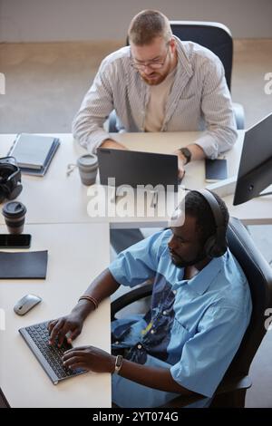 Two men sitting at office desk working on computers, collaborating on project. One colleague wearing headphones while typing Stock Photo