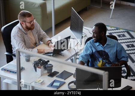 Two colleagues collaborating in a modern office, using laptops and technology to complete daily tasks. They are surrounded by desks, monitors, and office supplies Stock Photo