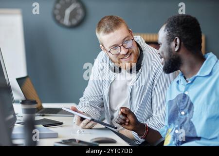 Two colleagues discussing project while using digital tablet and computer. One person, wearing glasses and headset, smiling and engaging in conversation Stock Photo