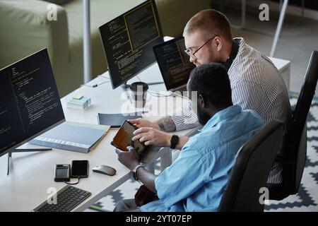 Two colleagues working together on software development in modern tech workspace, both focused on coding and using digital tools. One person is showing information on a tablet Stock Photo