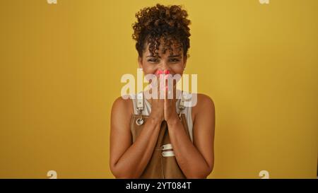 Young woman standing over isolated yellow background, wearing casual outfit and curly hair, with hands near her face and smiling playfully Stock Photo