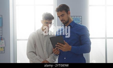 Two hispanic men discuss something on a digital tablet in a bright office, reflecting a modern workplace environment with a focus on collaboration and Stock Photo