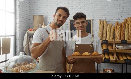 Two hispanic male bakers stand inside a bakery holding croissants, with one giving a thumbs-up and various bread items displayed on shelves in the bac Stock Photo