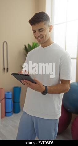 Young man using tablet in gym setting surrounded by exercise equipment, smiling and focused, captured in bright indoor environment with modern fitness Stock Photo