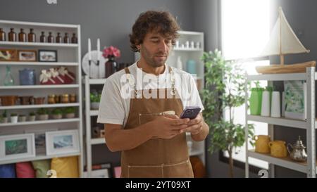 Young man in an apron standing inside a home decor store, using his phone, surrounded by various decorative items on shelves. Stock Photo