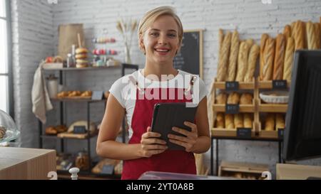 Young woman baker with short blonde hair holding a tablet in a well-lit bakery shop surrounded by bread and pastries Stock Photo