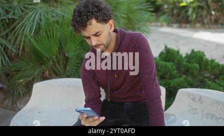 Young man with phone sits in urban park wearing maroon hoodie surrounded by greenery on a sunny day. Stock Photo