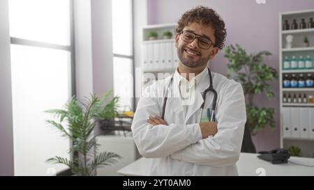 Hispanic male doctor with arms crossed smiling confidently inside a well-lit clinic office, showcasing his friendly and professional demeanor. Stock Photo
