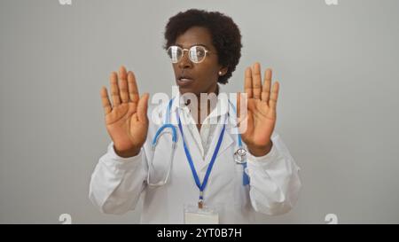 African american female doctor with curly hair and glasses makes a stop gesture with both hands, wearing a white coat with a stethoscope on an isolate Stock Photo