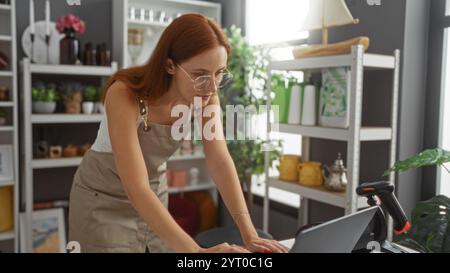 Woman working in a beautifully decorated home decor store, wearing glasses and an apron, surrounded by plants and decorative items, focusing intently Stock Photo