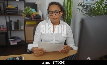 Woman working in an office, reading a document with a serious expression, surrounded by shelves and plants, illustrating a focused workplace environme Stock Photo