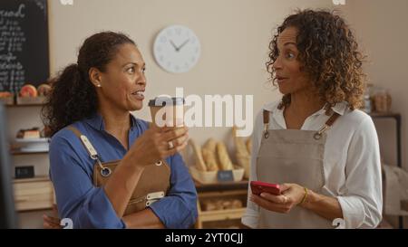 Women talking in a bakery shop with bread in the background, holding coffee and phone, wearing aprons, hispanic women partners engaged in conversation Stock Photo