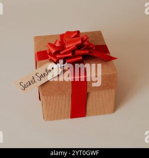 a gift box, tied with red ribbon and topped with a red ribbon bow, with a paper tag which reads the text secret santa, on a beige surface Stock Photo