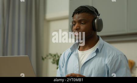 African American man user gamer guy male at home kitchen evening put on wireless headphones earphones with laptop computer listen music audio song Stock Photo