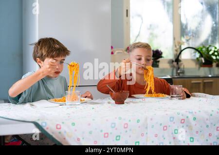 Two caucasian children enjoying a meal of spaghetti with tomato sauce, sitting at a table in a domestic kitchen Stock Photo