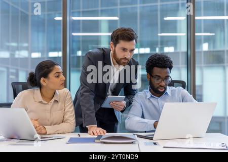 A diverse group of professional colleagues working together in an office space. They are focused and engaged, using a laptop and a tablet. This image highlights teamwork and collaboration. Stock Photo