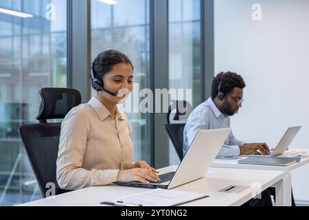 Two professionals in a modern office environment using laptops and headsets for communication. They appear focused and dedicated to their tasks, reflecting a productive workspace. Stock Photo