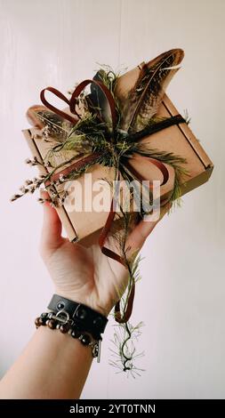 Craft packaging, package, box, parcel. Brown paper box decorated with dried herbs, feathers, ribbon, tapes in hand close up. Craft product, holiday Stock Photo