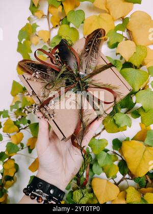 Craft packaging, package, box, parcel. Brown paper box decorated with dried herbs, feathers, ribbon, tapes in hand close up. Craft product, holiday Stock Photo