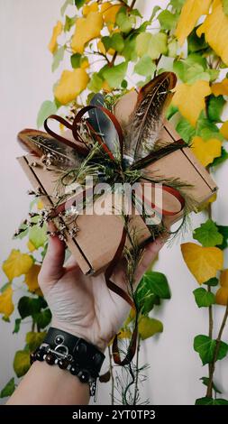 Craft packaging, package, box, parcel. Brown paper box decorated with dried herbs, feathers, ribbon, tapes in hand close up. Craft product, holiday Stock Photo