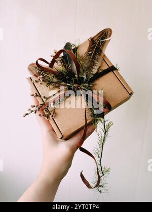 Craft packaging, package, box, parcel. Brown paper box decorated with dried herbs, feathers, ribbon, tapes in hand close up. Craft product, holiday Stock Photo