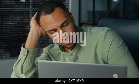 Tired lazy young african american businessman bored at work fall asleep at office desk sleepy overworked ethnic man employee sleeping at workplace Stock Photo