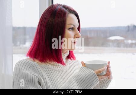 A woman with cherry red hair is holding a white cup of coffee. She is wearing a white sweater and is looking out the window. Closeup. Stock Photo