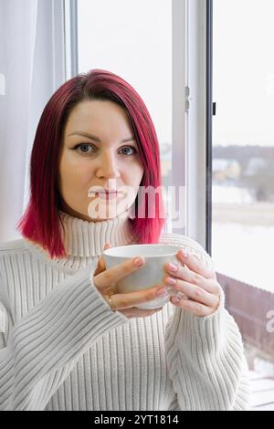 A woman with cherry red hair is holding a white cup of coffee. She is wearing a white sweater. Vertical crop. Closeup. Stock Photo